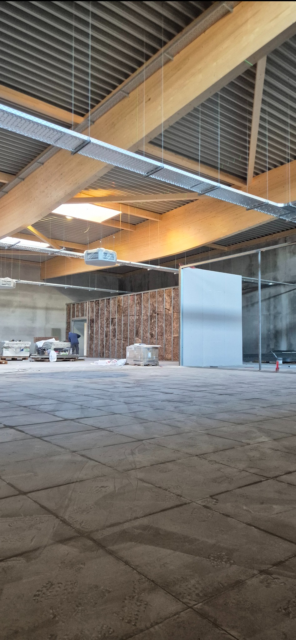 Wnętrze w trakcie prac wykończeniowych: widoczne drewniane belki stropowe, instalacje pod sufitem, ściany z izolacją i fragmenty ścian z płyt. W tle majster.