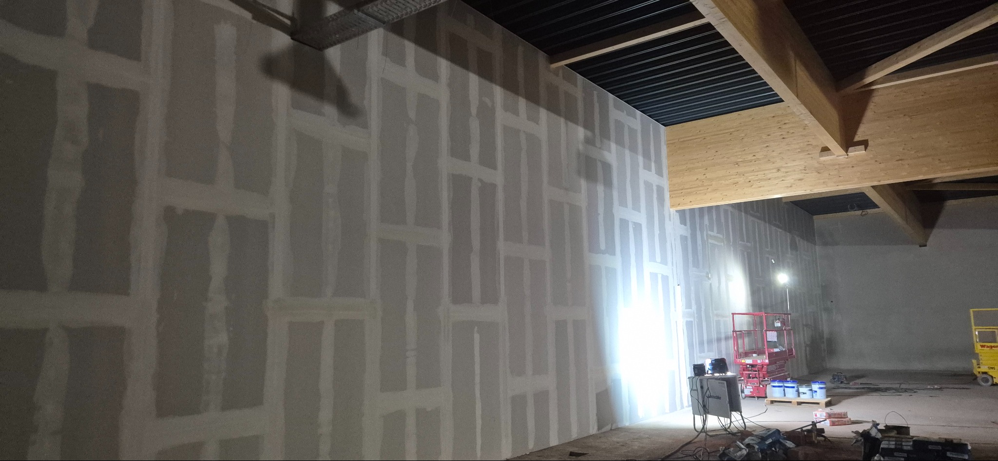 Ściana z płyt kartonowo-gipsowych w trakcie szpachlowania, widoczne łączenia. W tle podnośnik i sprzęt budowlany. Oświetlenie robocze.