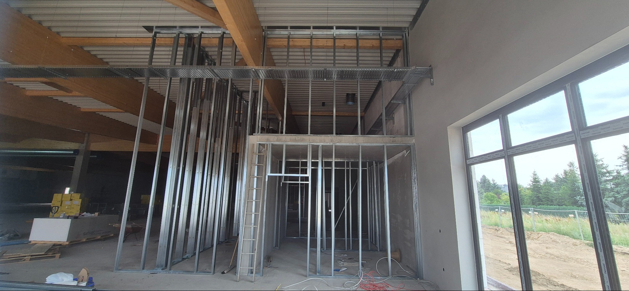 Wnętrze w trakcie prac: metalowa konstrukcja ścian działowych, drewniane belki stropowe, okno z widokiem na zieleń. Widoczna drabina i materiały budowlane.