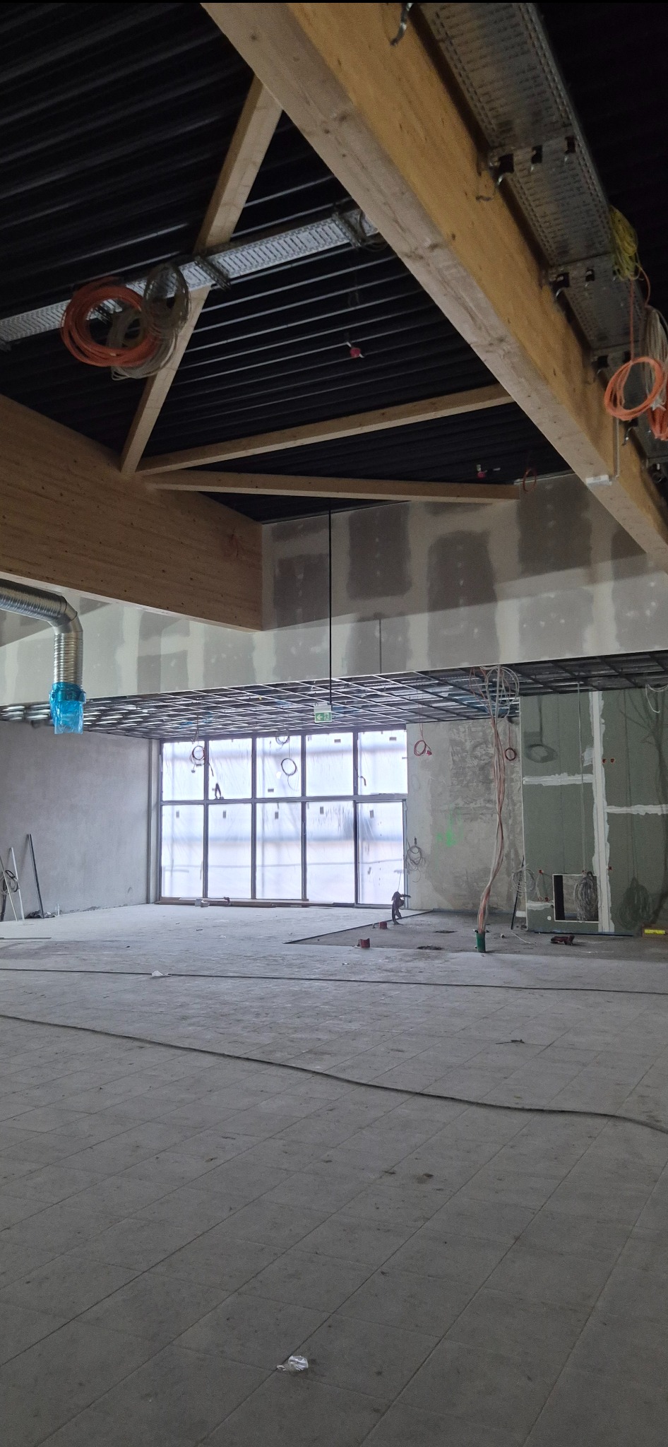 Wnętrze w trakcie prac wykończeniowych: widoczne drewniane belki stropowe, instalacje pod sufitem, ściany przygotowane pod malowanie, duże okno z folią ochronną.