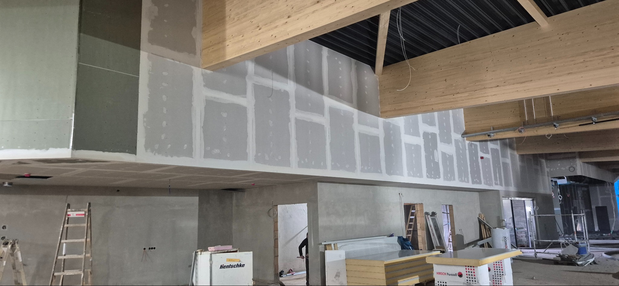 Wnętrze w trakcie prac wykończeniowych: ściany z płyt kartonowo-gipsowych, częściowo wyszpachlowane, drewniane belki stropowe, drabina oparta o ścianę. Widoczne materiały budowlane.