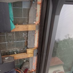 Yurii Shadura - Montaż okna dachowego z widoczną termoizolacją pianką montażową, folią paroizolacyjną i elementami konstrukcyjnymi dachu, częściowo zakryte siatką ochronną, z widocznym fragmentem dłoni w rękawicy...