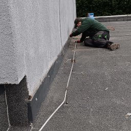 Yurii Shadura - Dekarz na dachu z papy termozgrzewalnej uszczelnia połączenie blachy przy ścianie budynku, widoczne mocowania instalacji odgromowej.