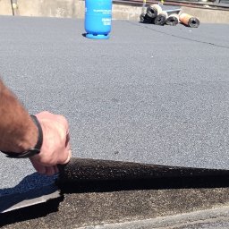 Yurii Shadura - Prace dekarskie: Ręka w czarnym zegarku odkleja starą warstwę papy z dachu garażu pokrytego szarą papą, w tle butla gazowa i rolki papy.