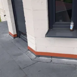 Płaski dach pokryty papą bitumiczną z widocznymi pęknięciami, detale obróbek blacharskich w kolorze ceglastym, okno z roletą zewnętrzną i butelką na parapecie, w tle samochód i przyczepa chłodnicza.