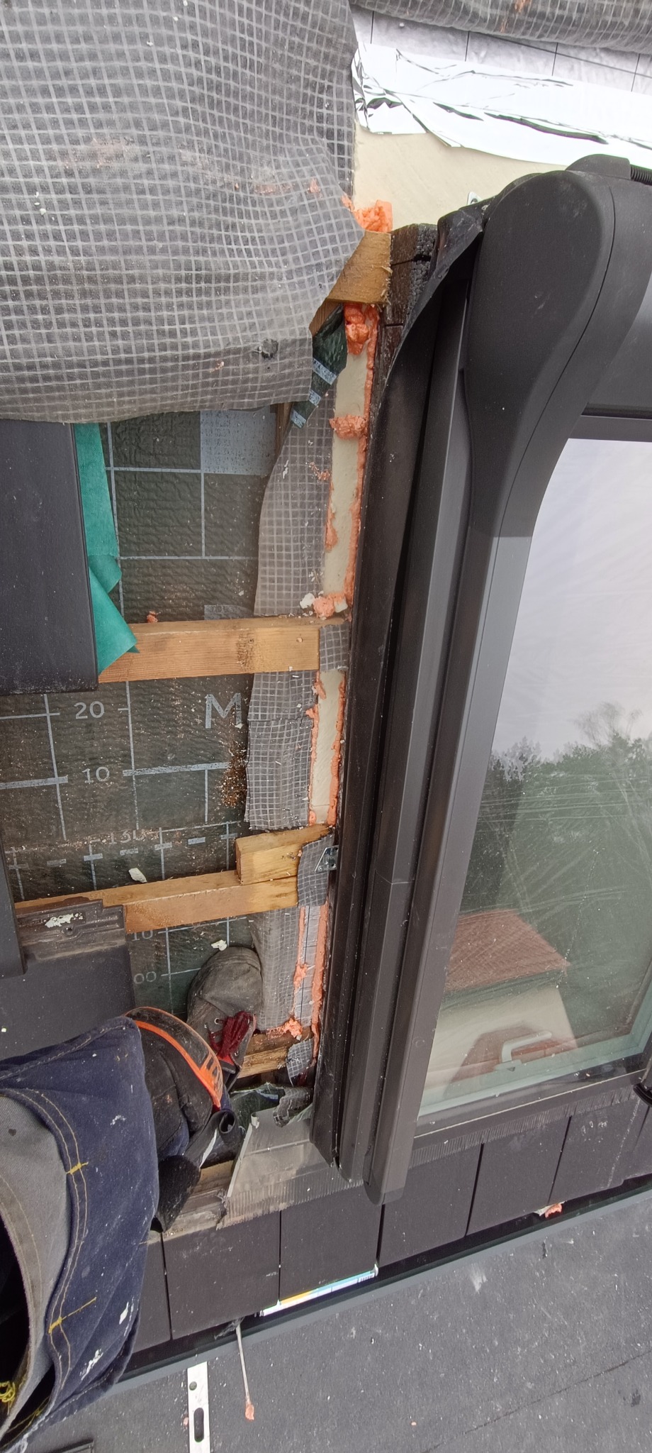 Montaż okna dachowego z widoczną termoizolacją pianką montażową, folią paroizolacyjną i elementami konstrukcyjnymi dachu, częściowo zakryte siatką ochronną, z widocznym fragmentem dłoni w rękawicy...