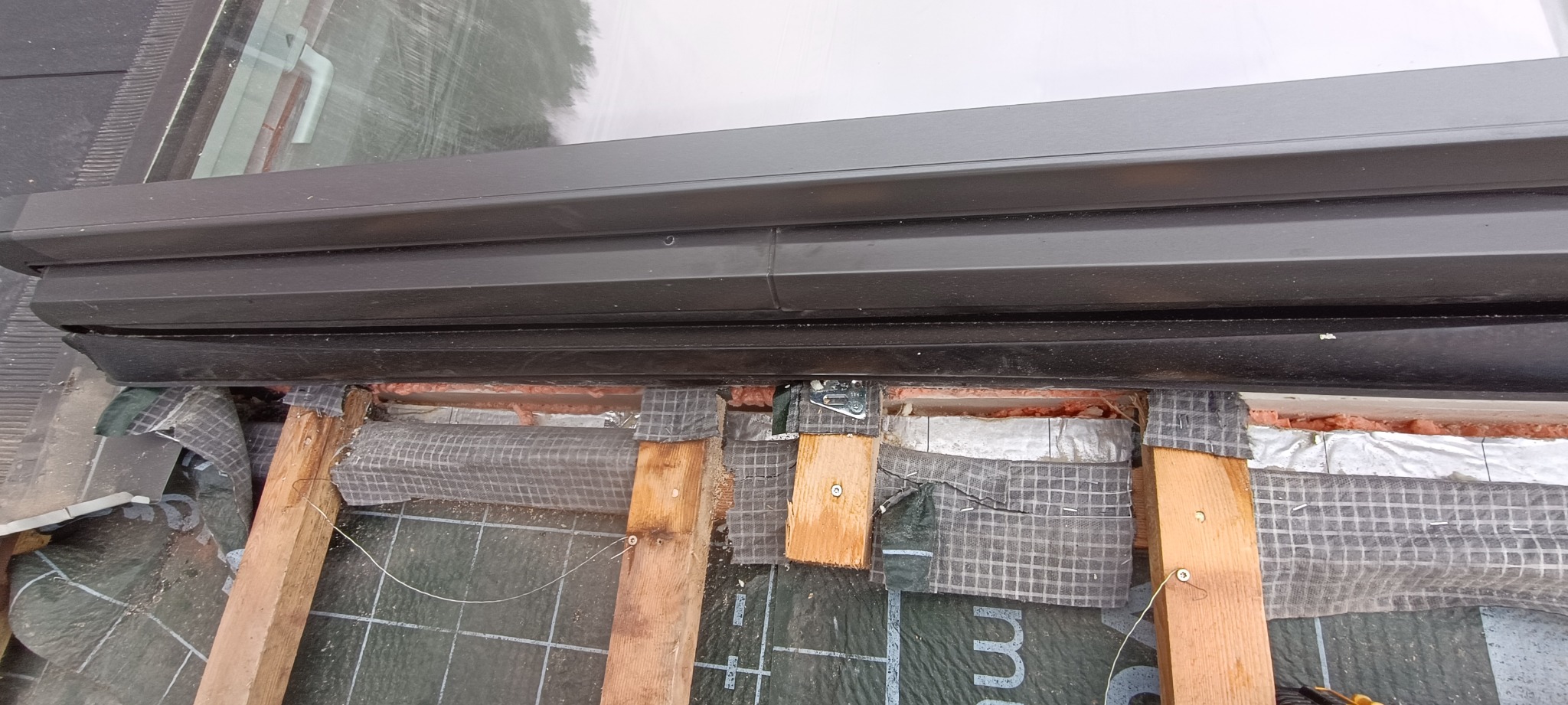 Szczegółowe ujęcie termoizolacji okna dachowego z widocznymi elementami konstrukcyjnymi i warstwami izolacyjnymi, zabezpieczonymi siatką i folią.