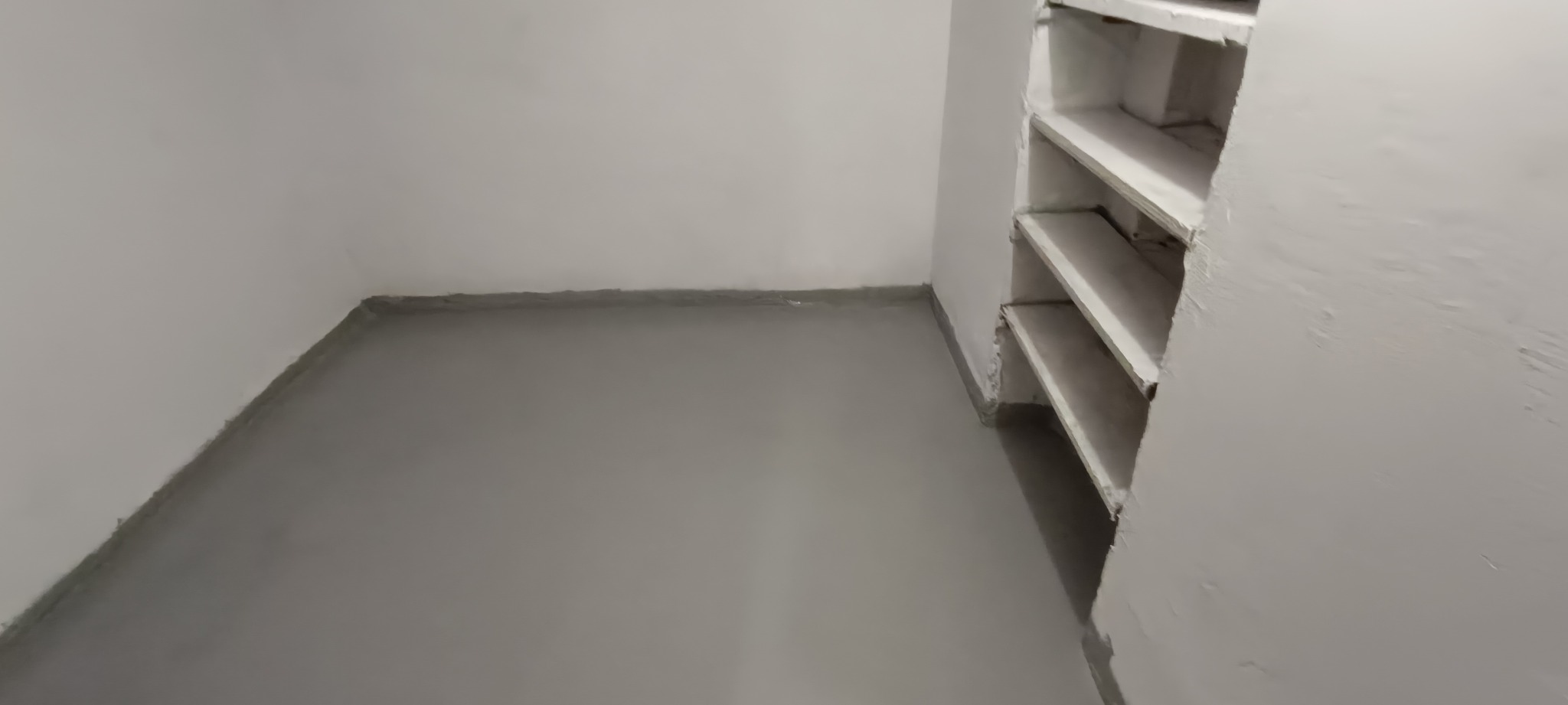 Szara podłoga żywiczna w piwnicy po wylaniu, widoczne ściany i fragment regału z półkami w rogu pomieszczenia.