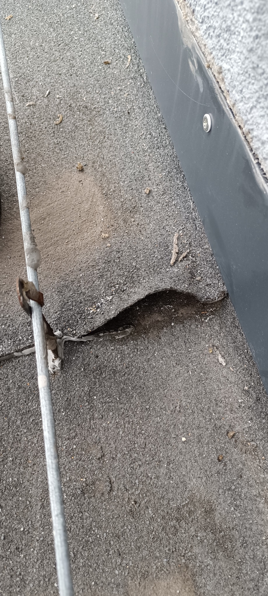 Uszkodzone pokrycie dachowe z papy z widocznym pęknięciem i odwarstwieniem od podłoża, obok metalowa rura odgromowa z zaciskiem.