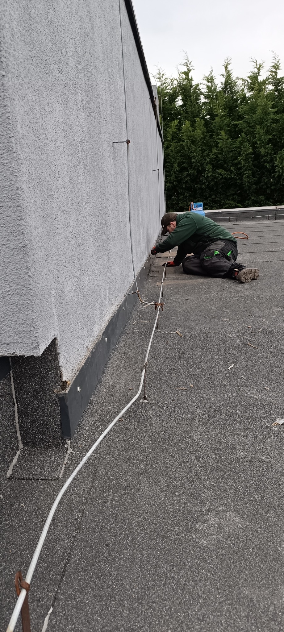 Dekarz na dachu z papy termozgrzewalnej uszczelnia połączenie blachy przy ścianie budynku, widoczne mocowania instalacji odgromowej.