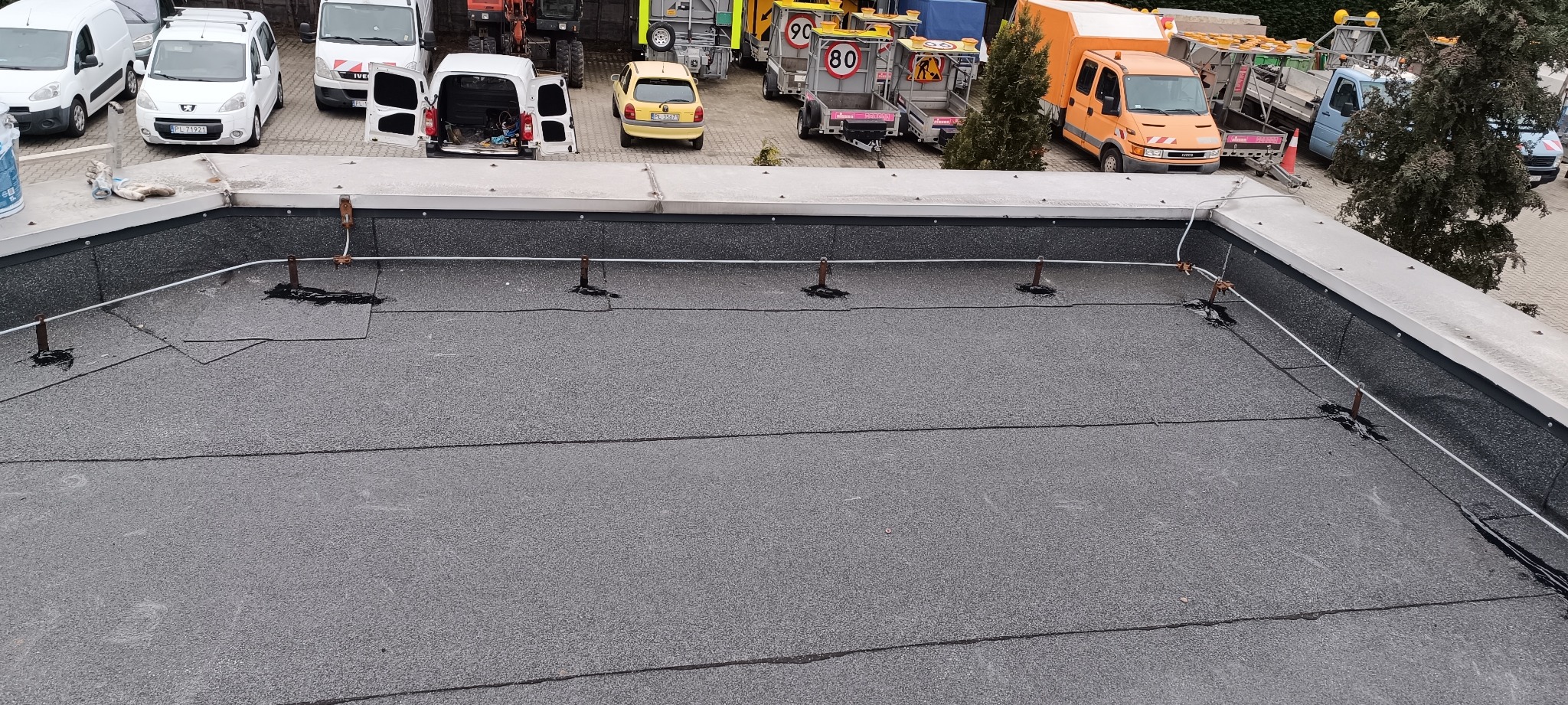 Płaski dach pokryty papą termozgrzewalną, z widocznymi elementami instalacji odgromowej i fragmentem naprawianego uszkodzenia, na tle zaparkowanych pojazdów.