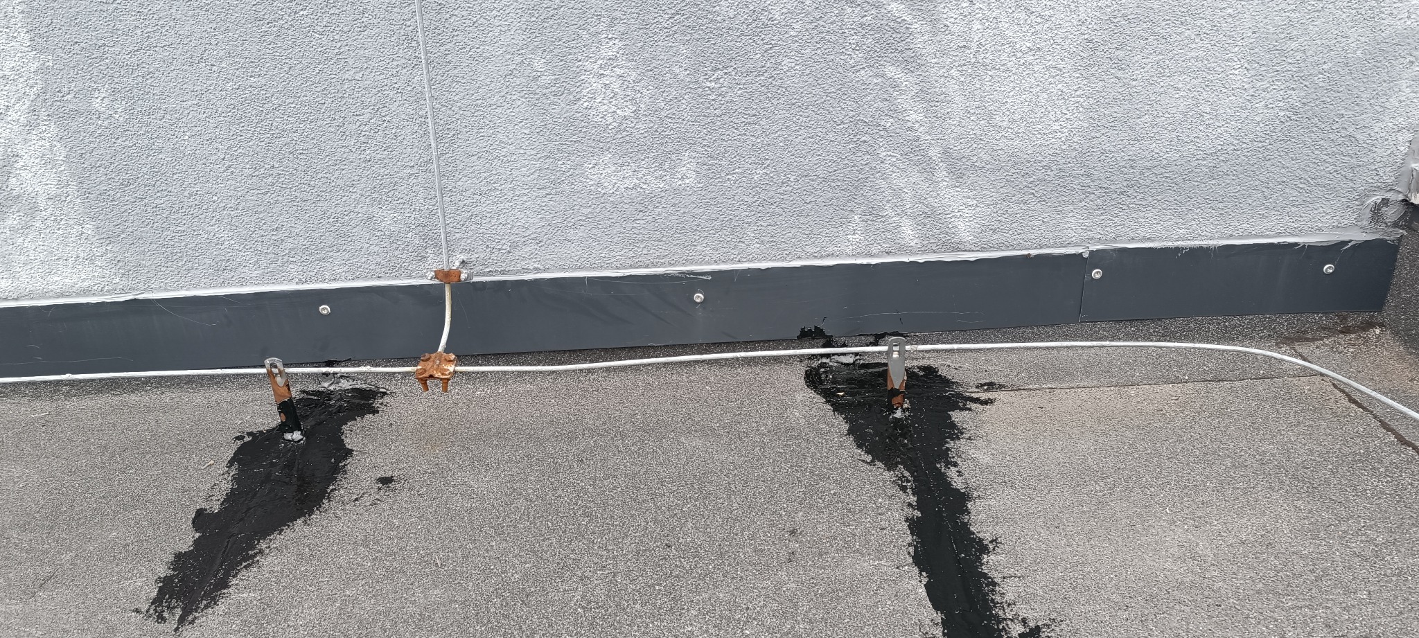 Płaski dach pokryty papą z widocznymi miejscami uszczelniania wokół metalowych elementów instalacji odgromowej, czarna masa bitumiczna, szara ściana budynku z widoczną fakturą tynku.