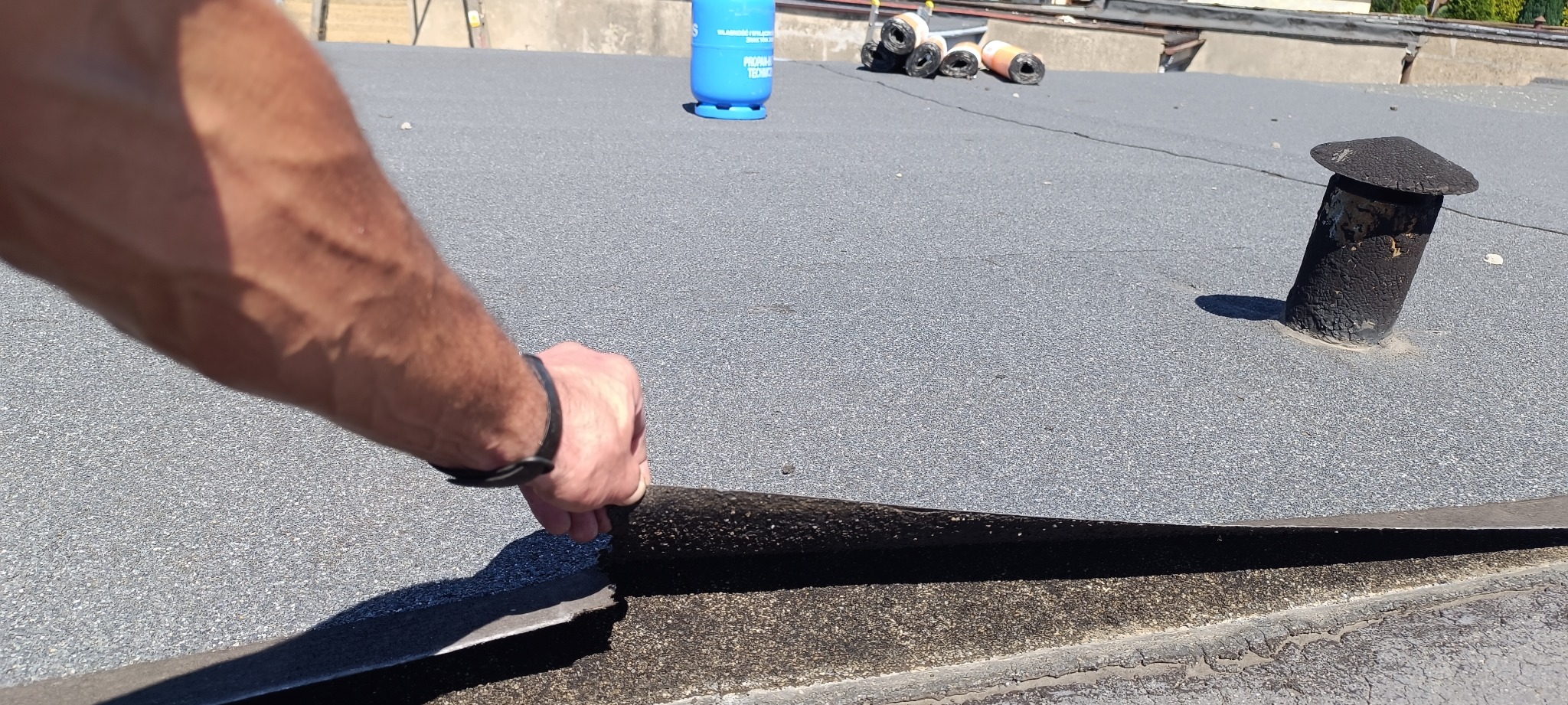 Prace dekarskie: Ręka w czarnym zegarku odkleja starą warstwę papy z dachu garażu pokrytego szarą papą, w tle butla gazowa i rolki papy.