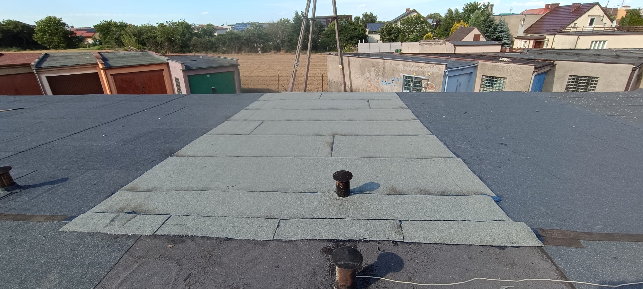 Fragment dachu garażu po naprawie, widoczne łatki z papy termozgrzewalnej na czarnej powierzchni, kominki wentylacyjne i rząd garaży w tle.