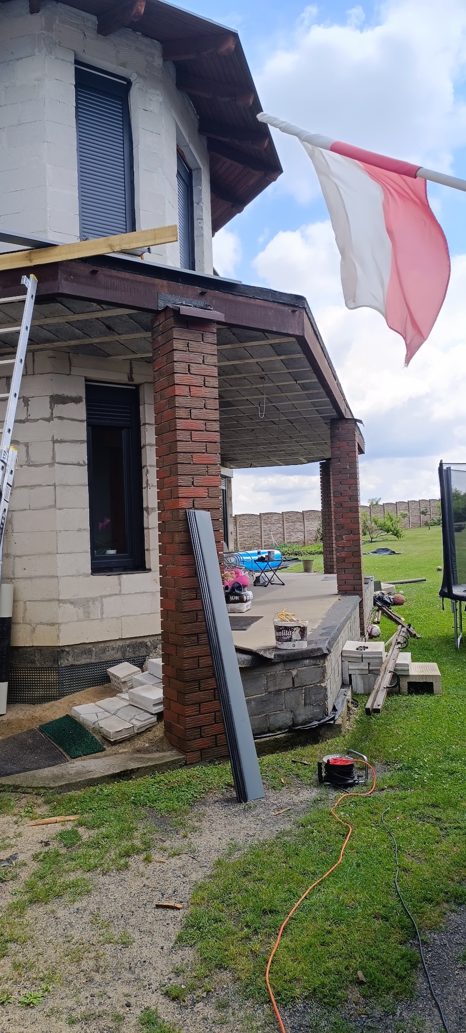 Dom w trakcie remontu z widocznymi elementami dachu i rynien, polska flaga powiewa na wietrze, elementy obróbki blacharskiej oparte o mur, rozłożone narzędzia i materiały budowlane na trawie, w tle...