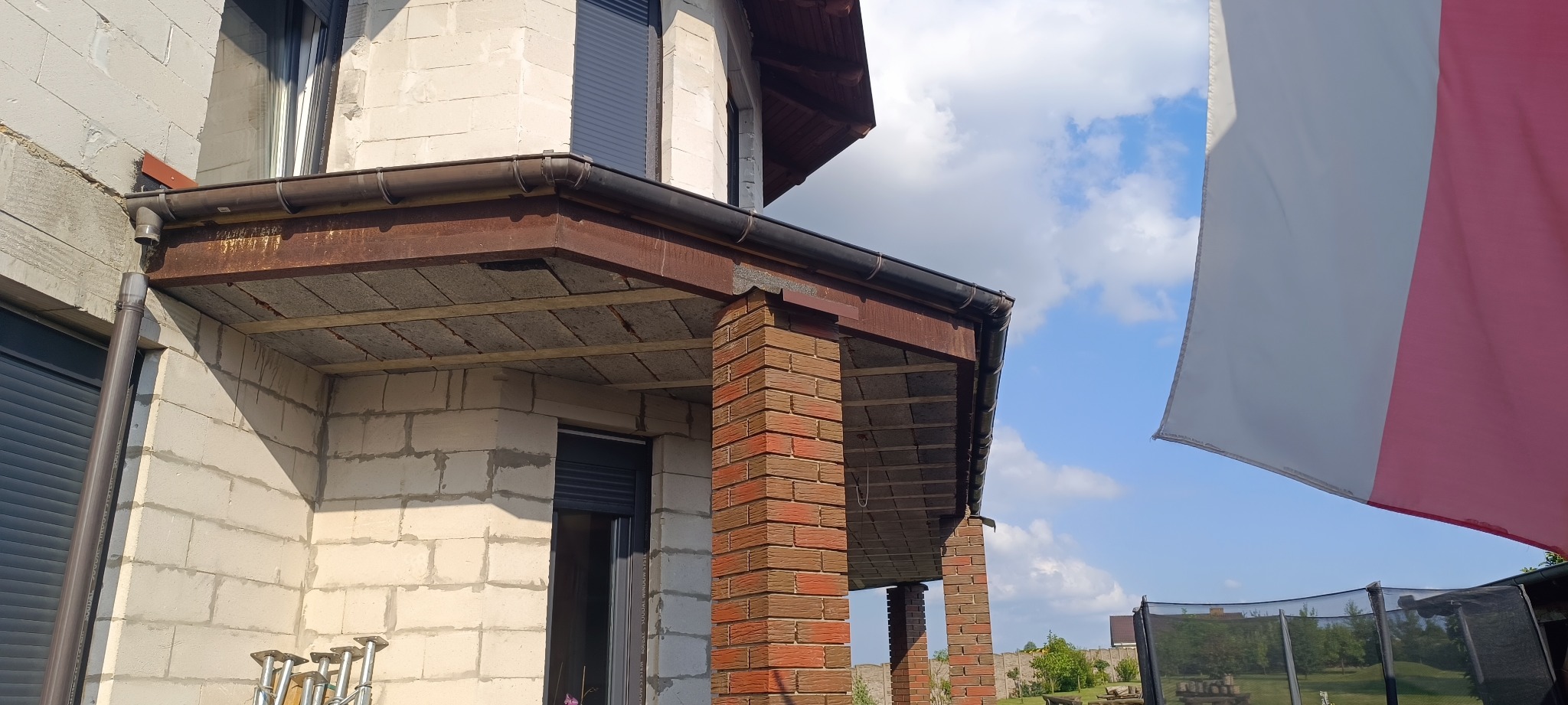 Fragment elewacji budynku w trakcie budowy z widocznymi rynnami, rurą spustową i fragmentem polskiej flagi w tle.