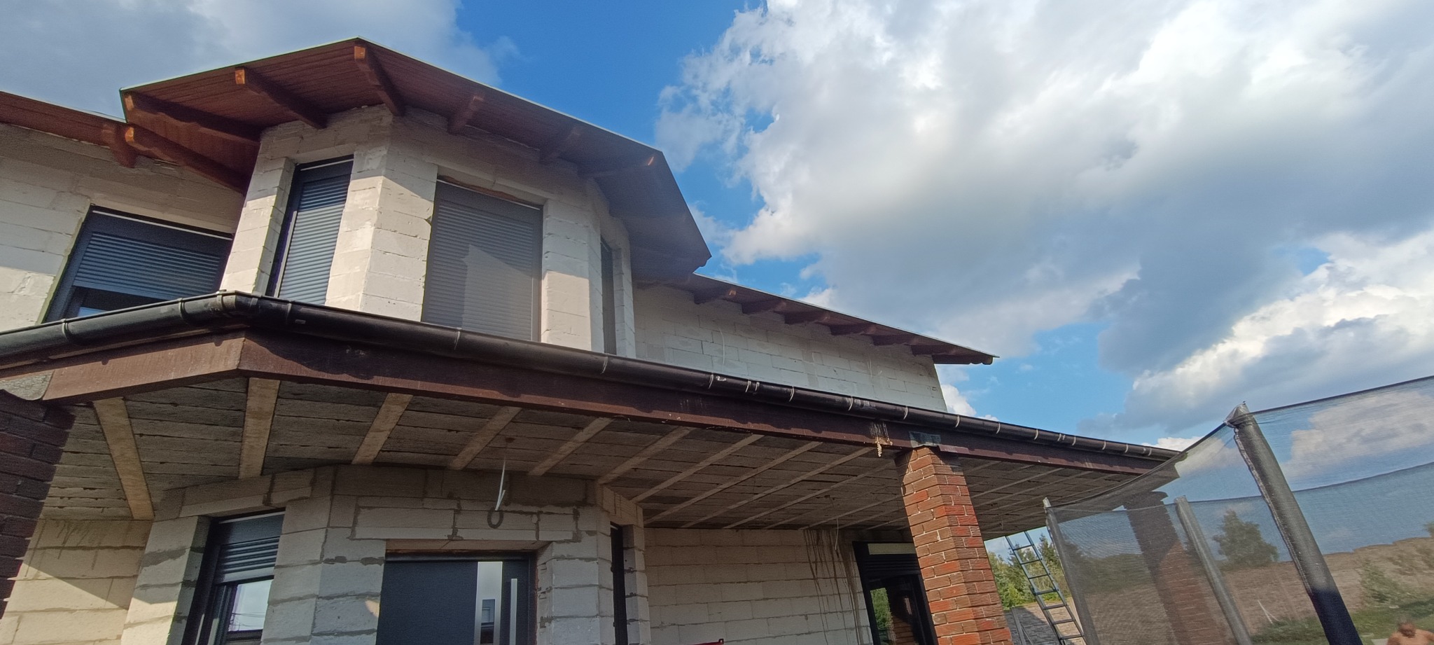 Niedokończony dom z białego bloczka z zamontowanymi, ciemnymi rynnami, widoczne drewniane elementy pod dachem i szare rolety w oknach, na tle błękitnego nieba z białymi chmurami.