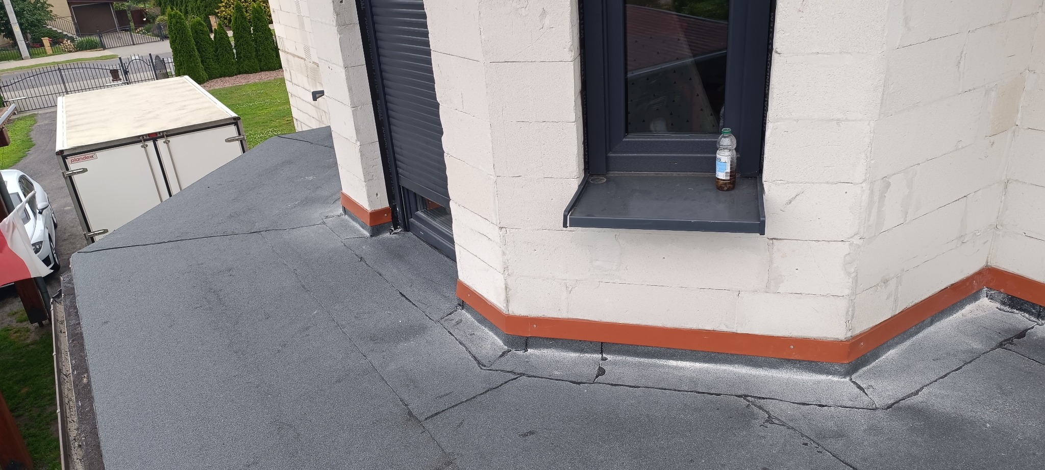 Płaski dach pokryty papą bitumiczną z widocznymi pęknięciami, detale obróbek blacharskich w kolorze ceglastym, okno z roletą zewnętrzną i butelką na parapecie, w tle samochód i przyczepa chłodnicza.