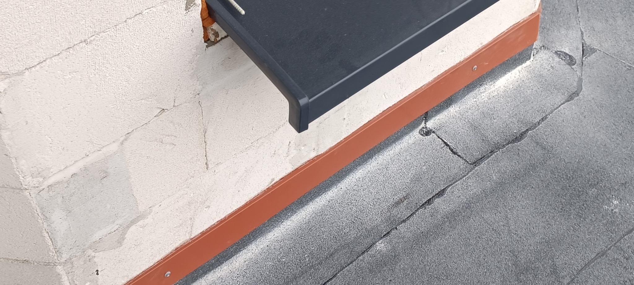 Fragment dachu z nową, grafitową obróbką blacharską okapową, pomarańczowa listwa rynnowa mocowana do ściany z białego betonu komórkowego, widoczne ślady uszczelniania pianką montażową.