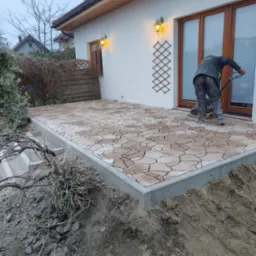 Układanie mozaiki brukowej na tarasie domu jednorodzinnego. Pracownik zagęszcza powierzchnię ubijarką w pomarańczowych rękawicach. Widoczne oświetlenie zewnętrzne.