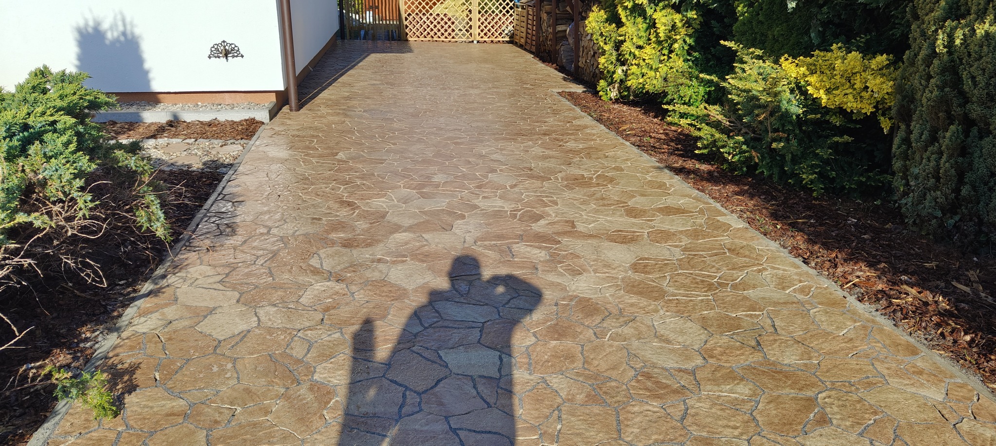Brązowa nawierzchnia z wzorem kamienia prowadząca do bramy, otoczona zielenią. Cień osoby na bruku. Widoczne elementy domu i dekoracje ścienne.