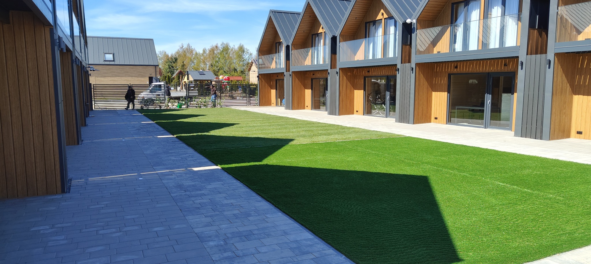 Nowoczesne domy szeregowe z kostką brukową i trawnikiem, widok z perspektywy chodnika. Na trawniku widoczne cienie, w tle osoby przy ogrodzeniu. Słoneczny dzień.