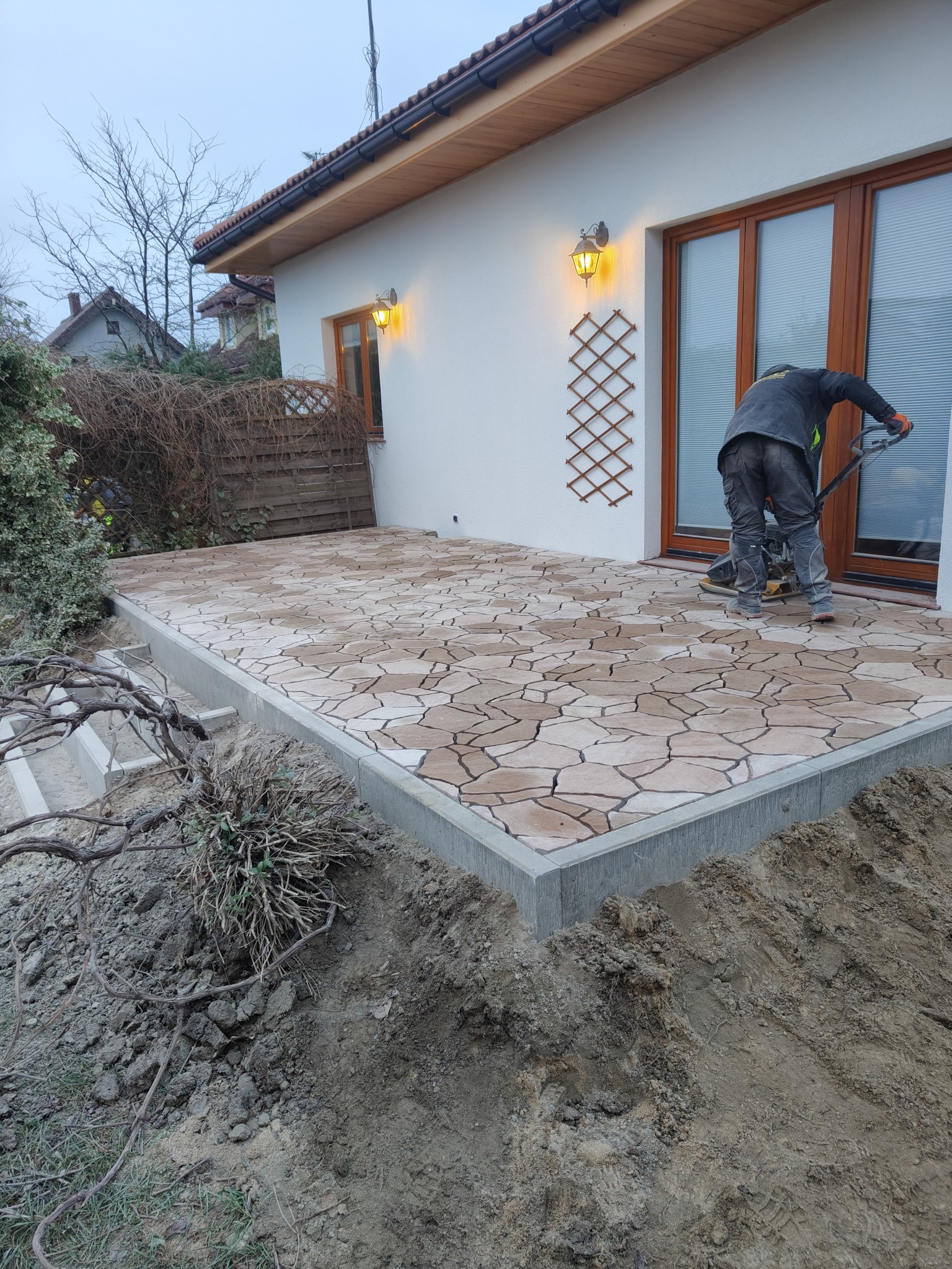 Układanie mozaiki brukowej na tarasie domu jednorodzinnego. Pracownik zagęszcza powierzchnię ubijarką w pomarańczowych rękawicach. Widoczne oświetlenie zewnętrzne.