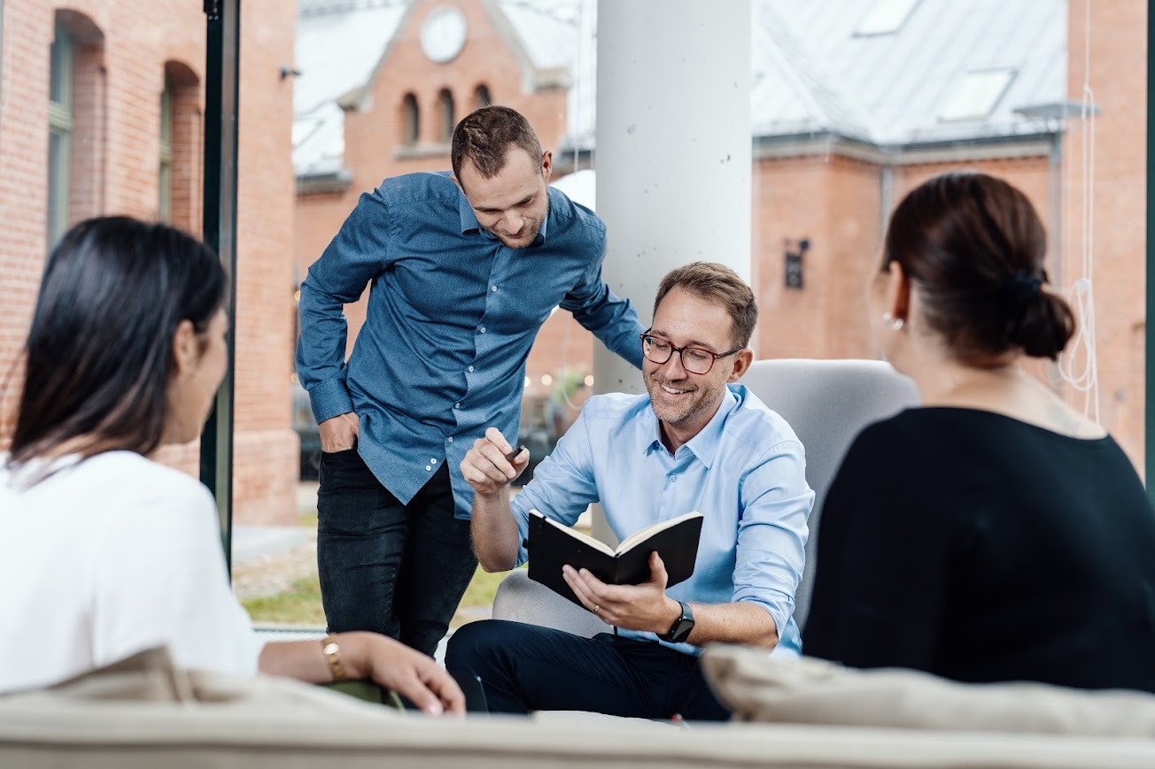 Konsultacja biznesowa w jasnym wnętrzu: mężczyzna w okularach omawia notatki z zespołem na tle ceglanego budynku widocznego przez okno.