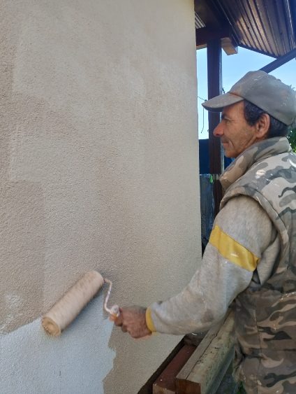 Mężczyzna w odzieży roboczej maluje zewnętrzną ścianę budynku wałkiem na kolor beżowy, widoczna nierówna faktura tynku.