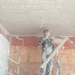 DAMIAN BUD - Pracownik na rusztowaniu aplikuje grubą warstwę tynku na sufit w surowym wnętrzu, widoczne cegły i nierówności.