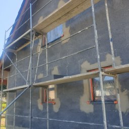 DAMIAN BUD - Dom w trakcie prac elewacyjnych: szare bloczki z widocznymi punktowymi zaprawami, rusztowanie metalowe, zabezpieczone okna folią.