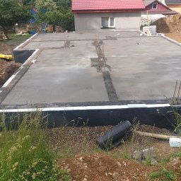 DAMIAN BUD - Wylana płyta fundamentowa domu jednorodzinnego, zabezpieczona izolacją pionową, z wystającymi prętami zbrojeniowymi, widok z poziomu gruntu.