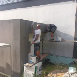 Dwóch pracowników na rusztowaniu tynkuje elewację budynku, widoczne puszki z tynkiem i narzędzia.