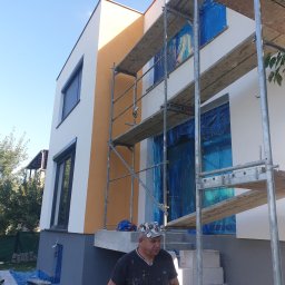 DAMIAN BUD - Dwukondygnacyjny dom z rusztowaniem, okna zabezpieczone folią, widoczny fragment elewacji w kolorze piaskowym, pracownik w ubraniu roboczym.