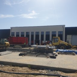 DAMIAN BUD - Plac budowy przed nowoczesnym budynkiem magazynowym z żółtą ładowarką kołową i czerwoną ciężarówką w słoneczny dzień.