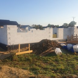 DAMIAN BUD - Wznoszenie murów parteru domu z białych bloczków, widoczna izolacja fundamentów, sterta ziemi i puste pojemniki po materiałach budowlanych na pierwszym planie.