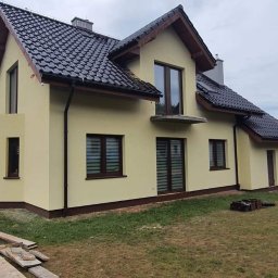 DAMIAN BUD - Żółta elewacja domu jednorodzinnego z ciemnobrązowymi oknami i dachem z dachówki ceramicznej, widoczne deski na trawniku.