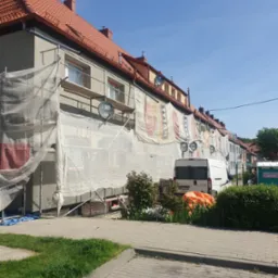 Ocieplanie elewacji szeregu domów jednorodzinnych, osłoniętych siatką budowlaną i rusztowaniami, z widocznym samochodem dostawczym i częściowo wykończoną elewacją.