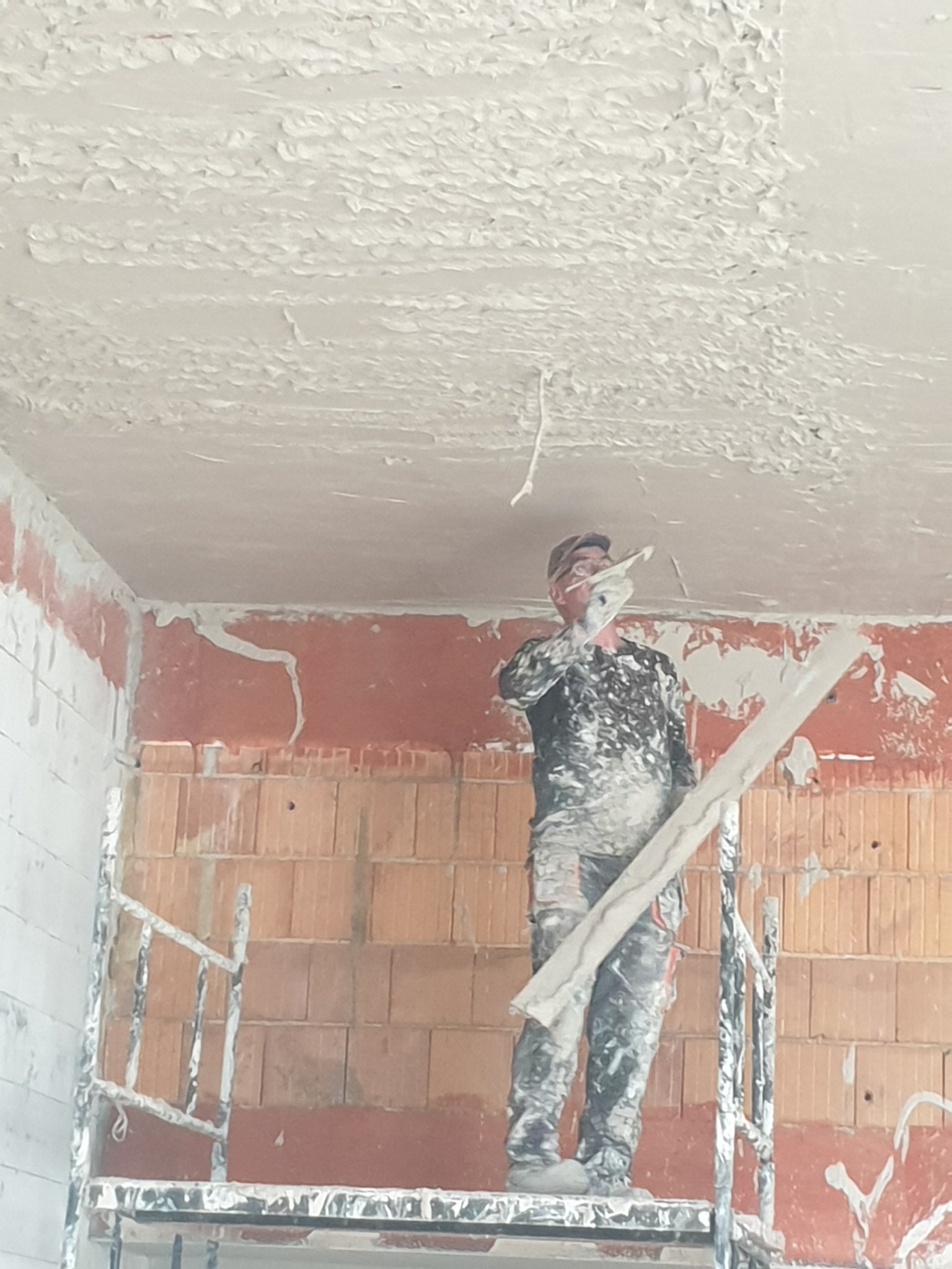Pracownik na rusztowaniu aplikuje grubą warstwę tynku na sufit w surowym wnętrzu, widoczne cegły i nierówności.