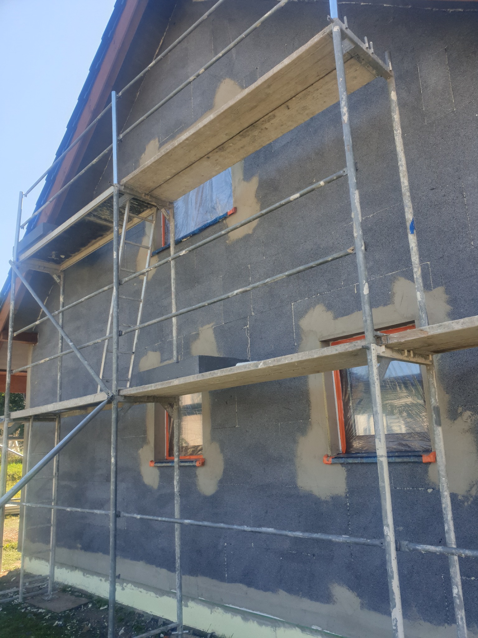 Dom w trakcie prac elewacyjnych: szare bloczki z widocznymi punktowymi zaprawami, rusztowanie metalowe, zabezpieczone okna folią.