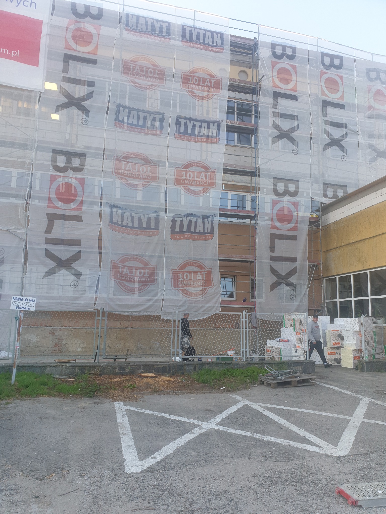 Budynek w trakcie prac, osłonięty siatką z reklamami, widoczne rusztowania i materiały izolacyjne, dwóch mężczyzn przechodzi obok.
