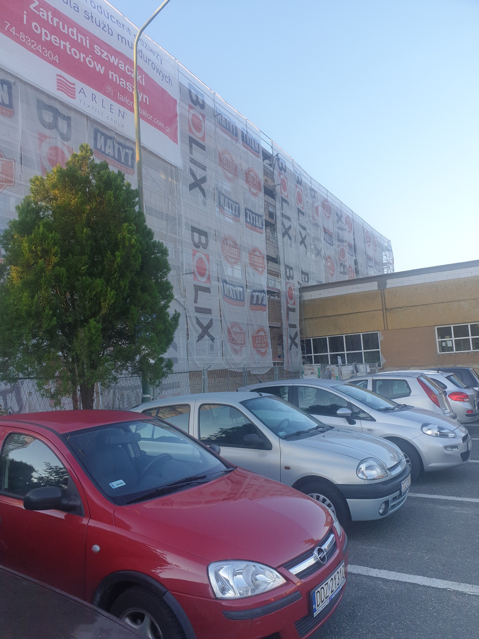 Budynek w rusztowaniach osłonięty siatką z reklamami firm Bolix i Arlen Textile Group, częściowo widoczny za zielonym drzewem na parkingu z zaparkowanymi samochodami.