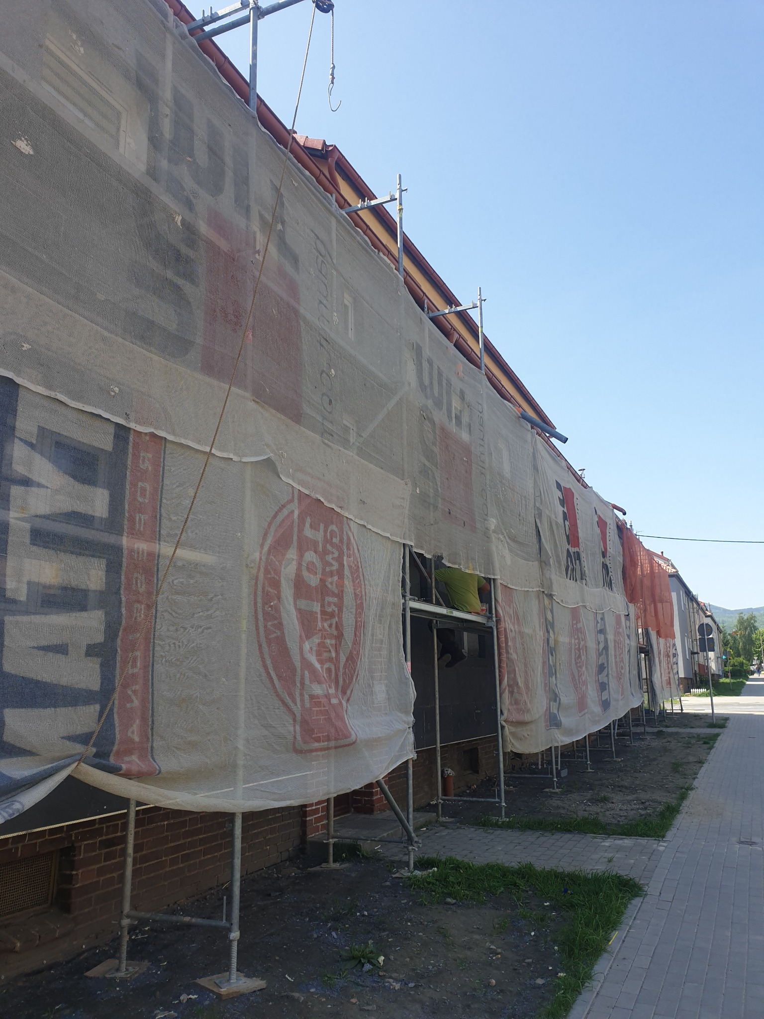 Budynek w trakcie remontu elewacji, osłonięty siatką z reklamami, widoczne rusztowania i fragment pracującego robotnika.