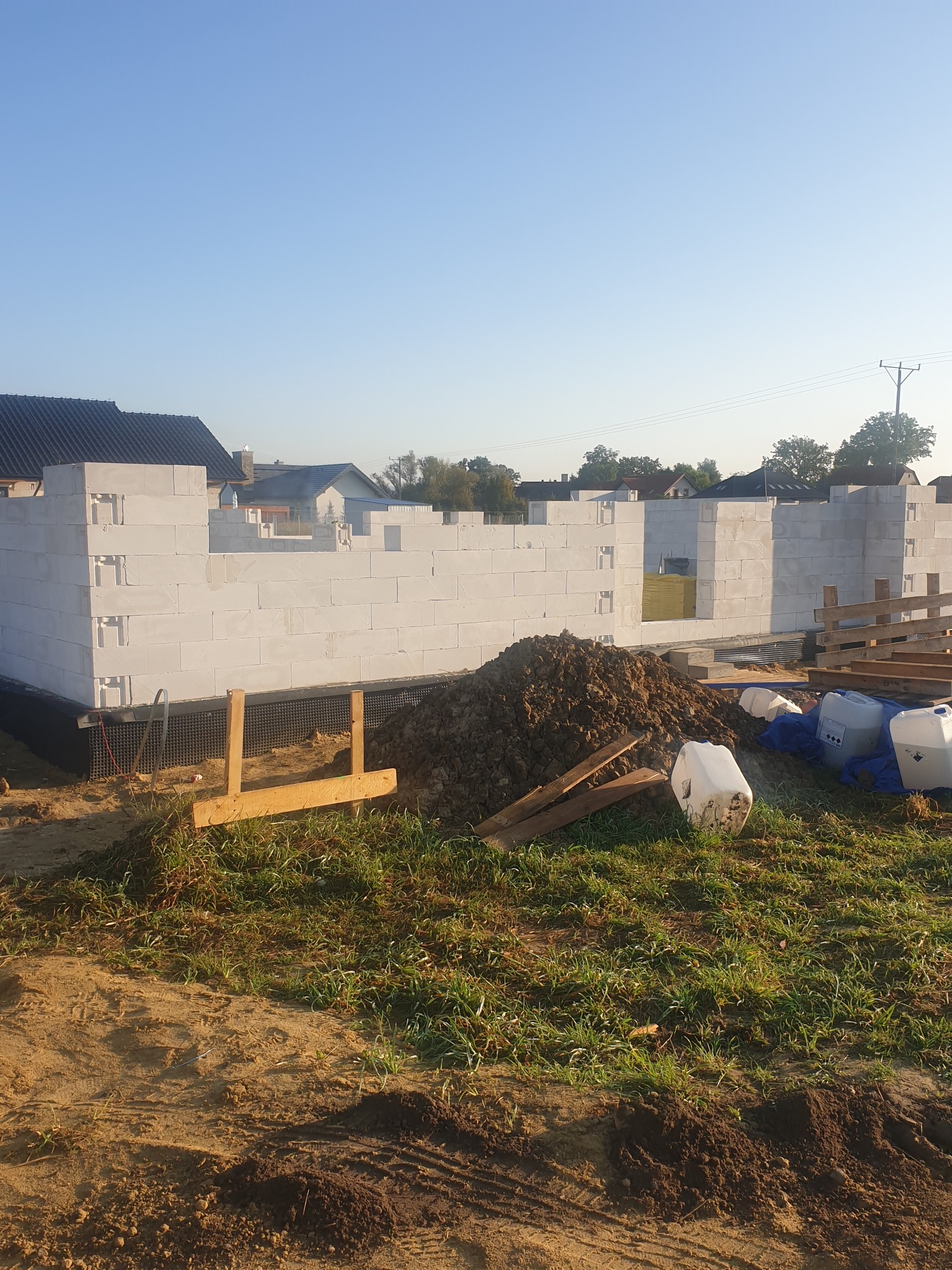 Wznoszone ściany budynku z bloczków betonu komórkowego Ytong na fundamencie z hydroizolacją, widoczne zapasy materiałów budowlanych i świeżo nawieziona ziemia.