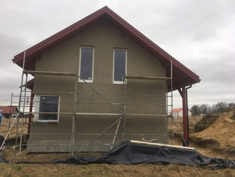 Dom jednorodzinny w trakcie prac elewacyjnych, widoczne rusztowanie i świeżo nałożony tynk w kolorze beżowym, czerwona podbitka dachowa