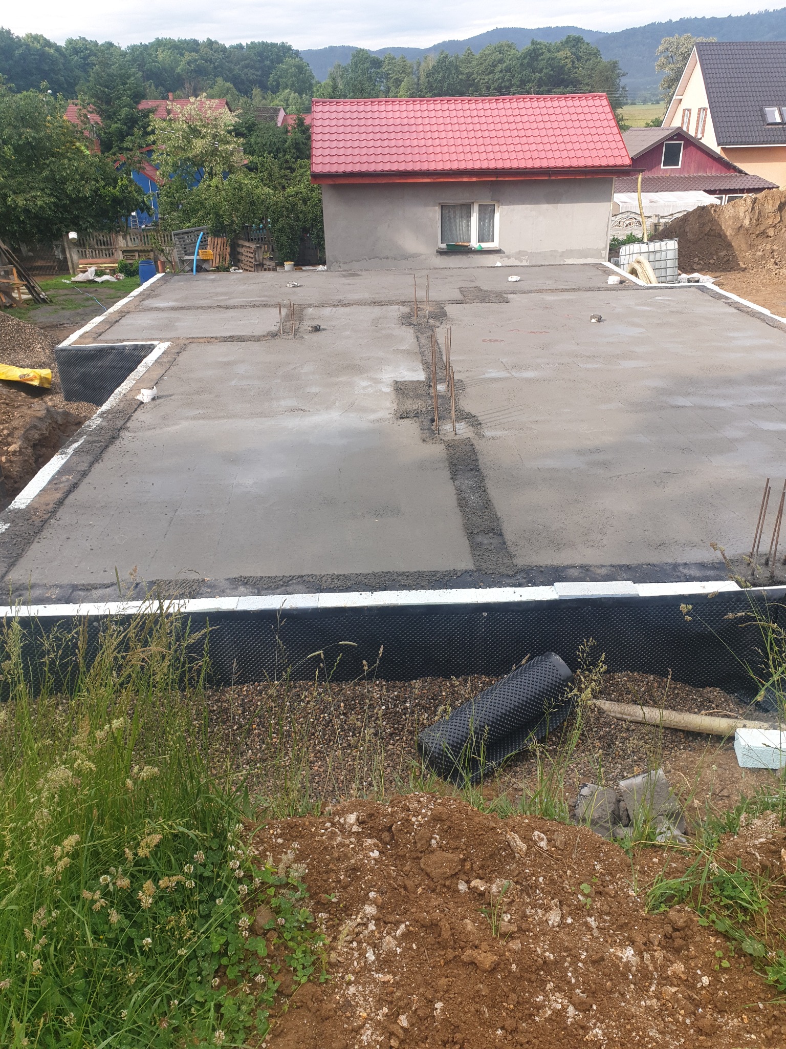 Wylana płyta fundamentowa domu jednorodzinnego, zabezpieczona izolacją pionową, z wystającymi prętami zbrojeniowymi, widok z poziomu gruntu.