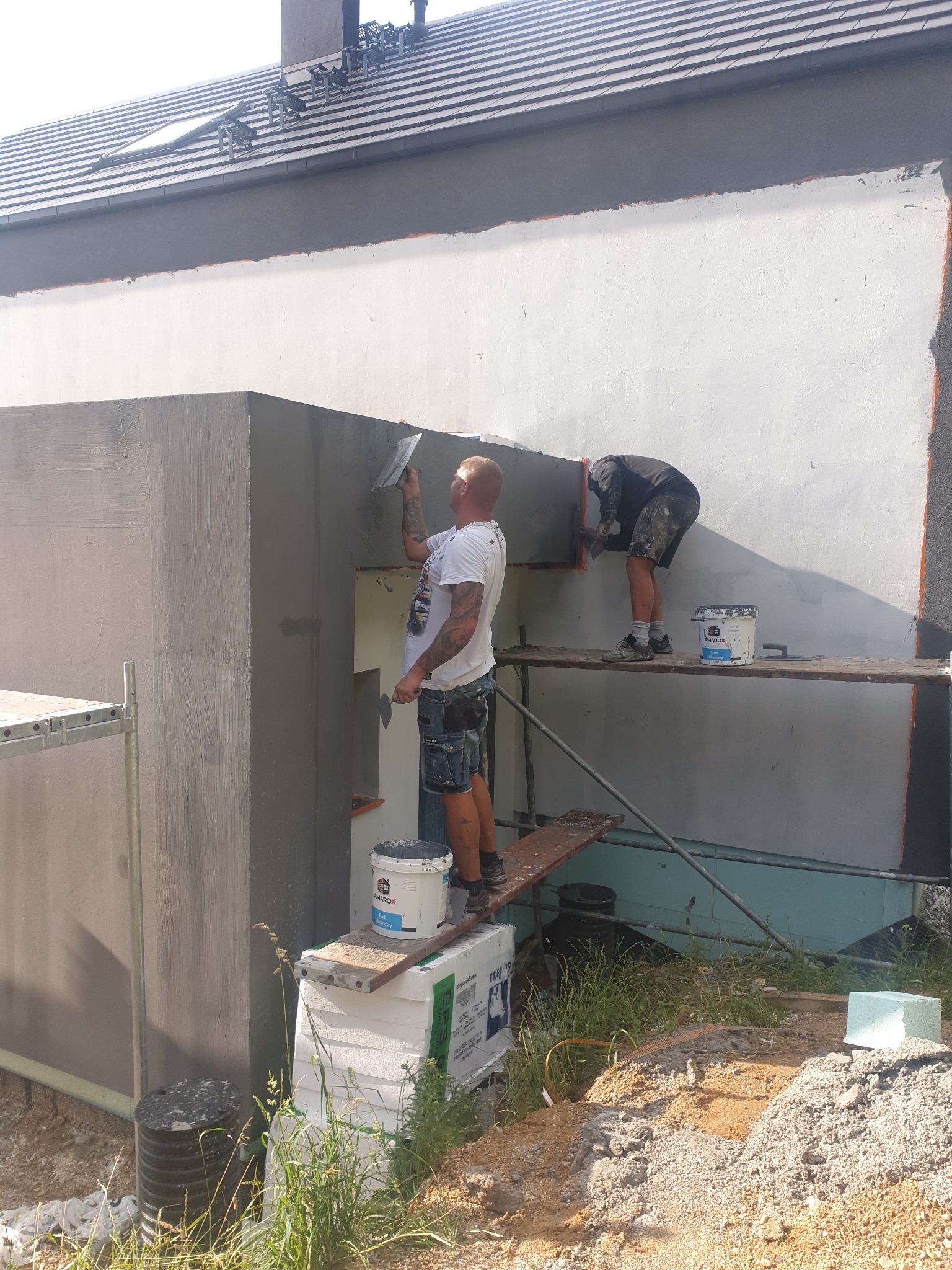 Dwóch pracowników na rusztowaniu tynkuje elewację budynku, widoczne puszki z tynkiem i narzędzia.