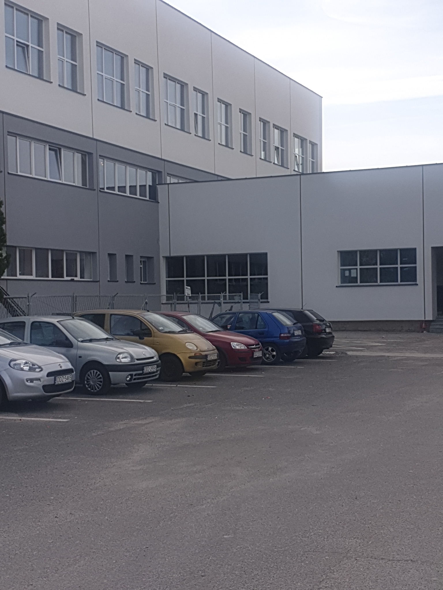 Szary, dwukondygnacyjny budynek biurowy z rzędem zaparkowanych samochodów osobowych przed nim, widok z poziomu parkingu.