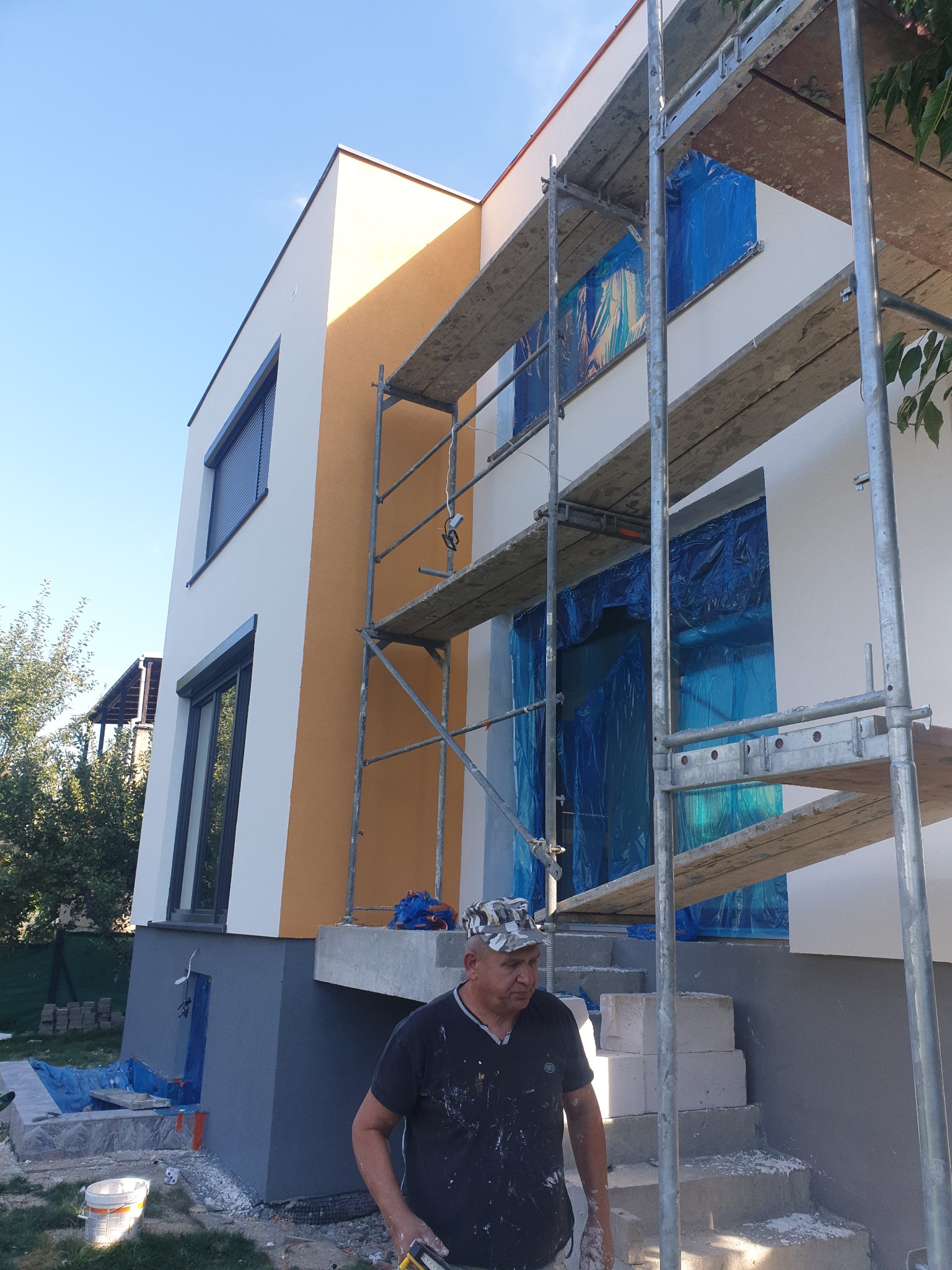 Dwukondygnacyjny dom z rusztowaniem, okna zabezpieczone folią, widoczny fragment elewacji w kolorze piaskowym, pracownik w ubraniu roboczym.