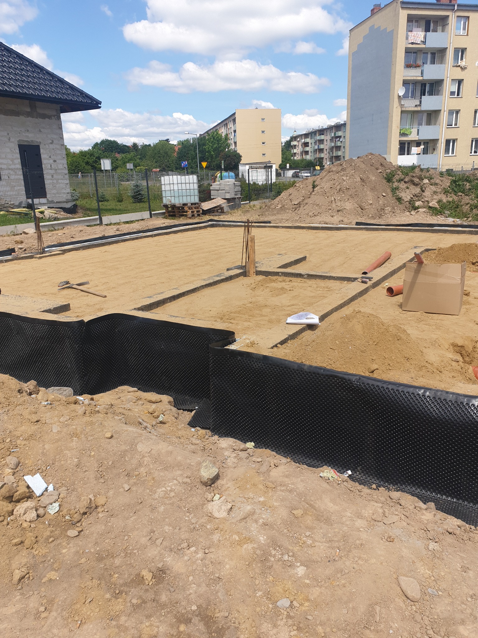 Przygotowanie terenu pod fundamenty domu jednorodzinnego z widoczną izolacją pionową ścian fundamentowych, piaskiem zasypanym wewnątrz obrysu fundamentów i widoczną okoliczną zabudową wielorodzinną...